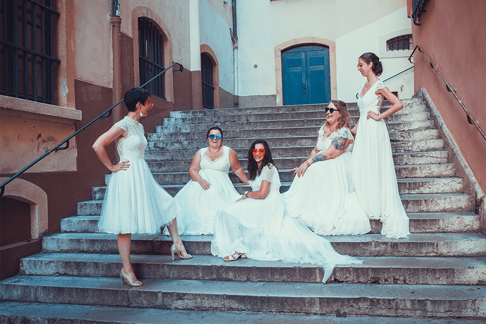 Cinq femmes en robes de mariée dont deux grandes tailles assises sur des escaliers