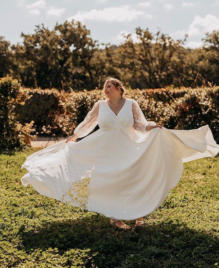 Mariée en robe sur-mesure pour femme ronde tournant joyeusement dans un jardin, manches longues en dentelle